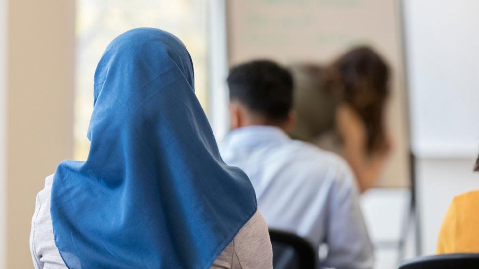 A stock image of a girl wearing a blue headscarf. She is sitting down in a classroom, facing with her back towards the camera.
