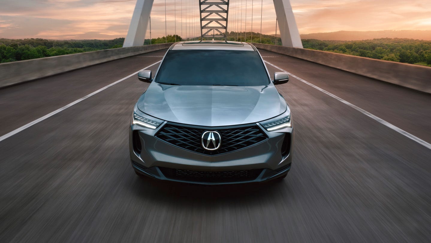 front view of a silver suv driving on a bridge at sunset, with a steel arch structure and forested landscape in the background.