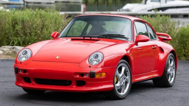 Red sports car parked near water, with a scenic background of trees and houses.