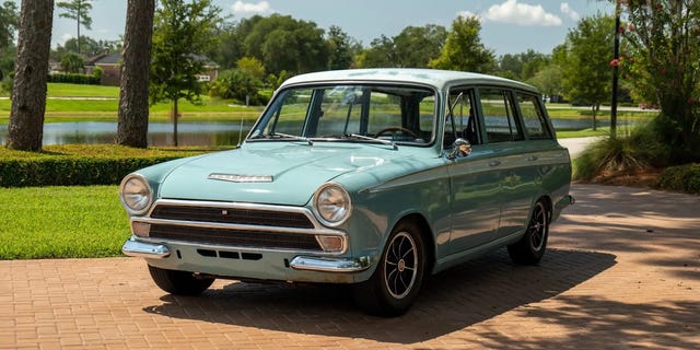 Vintage light blue station wagon parked on a brick driveway with trees and a lake in the background.